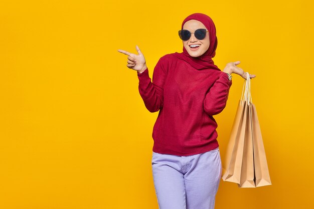 Mulher asiática jovem e alegre segurando sacolas de compras e apontando o dedo para o espaço da cópia isolado em fundo amarelo