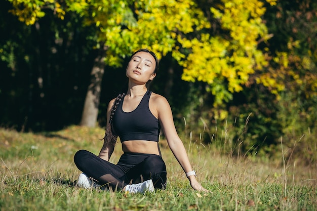 Mulher asiática jovem e bonita esportiva meditando no parque, sentada em pose de lótus praticando tapete de ioga, zen. relaxa ao ar livre na natureza pela manhã Conceito de estilo de vida saudável, relaxe, calma, meditação