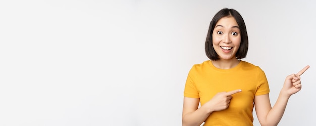 Mulher asiática jovem espantada mostrando propaganda de lado apontando dedos para o logotipo da marca de texto de promoção em pé feliz contra o fundo branco