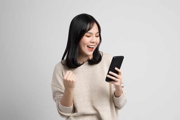 Mulher asiática jovem feliz olhando para a tela do smartphone em pé sobre fundo branco isolado. Ela muito feliz.