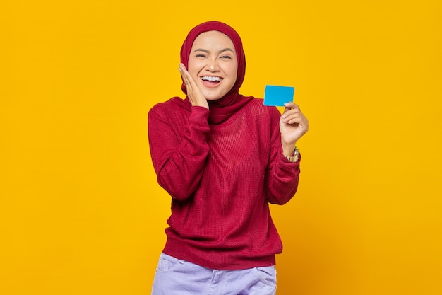 Mulher asiática jovem feliz segurando um cartão de crédito e tocando as bochechas em fundo amarelo