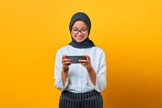 Mulher asiática jovem feliz usando o celular para jogar no fundo amarelo