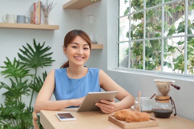 Mulher asiática jovem feliz usando tablet e tomando café pela manhã