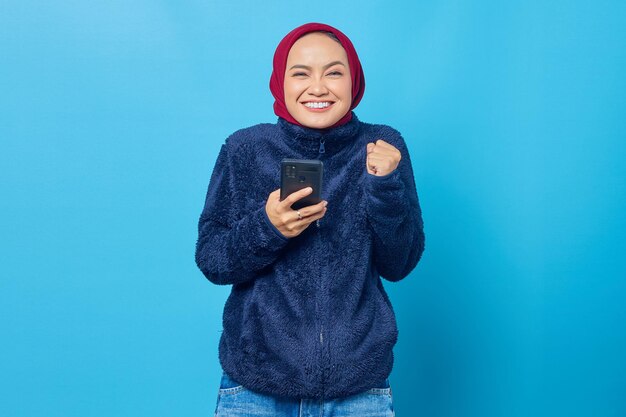 Mulher asiática jovem feliz usando telefone celular com gesto de mão bem-sucedido em fundo azul