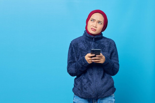 Mulher asiática jovem pensativa usando smartphone e olhando para cima isolada sobre fundo azul