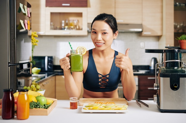 Mulher asiática jovem positiva com uma xícara de suco verde mostrando o polegar para cima e sorrindo para a câmera