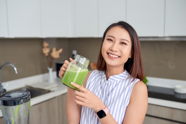 Mulher asiática jovem sorridente segurando suco verde na cozinha em casa.