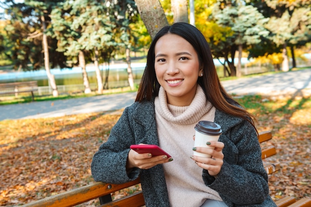 Mulher asiática jovem sorridente, vestindo um casaco, sentada em um banco do parque, usando telefone celular