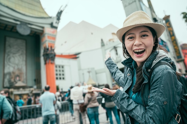 mulher asiática jovem viajante sorrindo com expressão satisfeita mostrando o palácio do filme com as duas mãos. excitada turista feminina boca aberta larga rindo compartilhando rosto câmera teatro chinês hollywood street