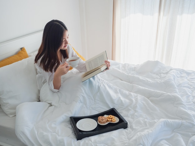 Mulher asiática lendo livro e tomando café na cama de manhã