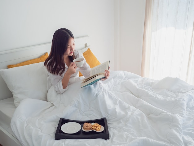 Mulher asiática lendo livro e tomando café na cama de manhã