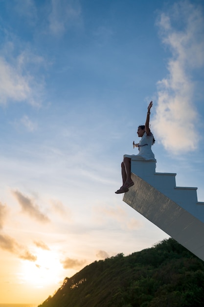 Mulher asiática levanta os braços, sente-se no topo da escada de madeira com o céu do sol ao fundo, vitória, conceito de sucesso