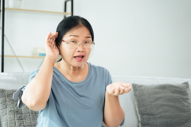 Mulher asiática madura com surdez e deficiência auditiva que tem problemas de audição segura a mão dele sobre o ouvido e escuta com atenção, com problemas de audição.