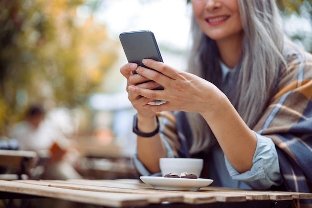 Mulher asiática madura feliz escreve a mensagem no telefone, sentado à mesa no terraço do café ao ar livre