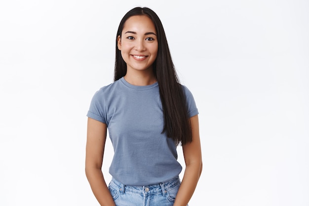 Mulher asiática moderna bonita com longos cabelos escuros, sorrindo feliz e olhando a câmera, pose casual em pé, pensando na próxima festa de Natal, expressar emoções positivas e alegres, parede branca