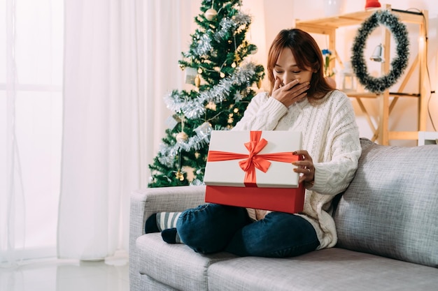 mulher asiática muito feliz está cobrindo a boca de surpresa por receber um presente valioso na manhã de natal em um interior de sala de estar em casa festiva.