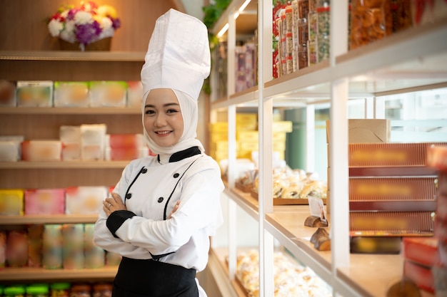 Mulher asiática muito muçulmana com uniforme de chef usando hijab cruzou o braço na frente da loja. proprietário de uma pequena empresa muçulmana