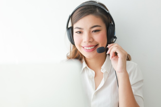 Mulher asiática na camisa branca falar com fone de ouvido e usando o laptop, o sorriso e o conceito de operador de rosto feliz