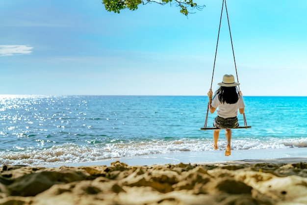 Mulher asiática no estilo casual usar chapéu balançar os balanços na praia de areia e olhando o paraíso tropical lindo mar e céu no pôr do sol. Férias de verão. Vibes do verão.