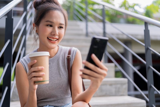 Foto mulher asiática nômade digital despreocupada mão segura xícara de café enquanto usa smartphone conversando com cliente remoto enquanto está sentado na escada do parque público cidade urbana estilo de vida moderno trabalhando