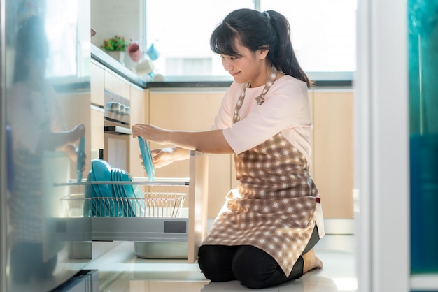Mulher asiática nova atrativa que carrega a máquina de lavar louça em armários na cozinha ao fazer a limpeza em casa durante ficar em casa usando o tempo livre sobre sua rotina diária de limpeza.