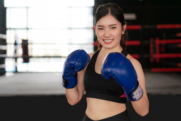 Mulher asiática nova da aptidão do encaixotamento que sorri feliz vestindo luvas de boxe azuis