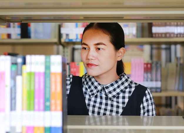 Mulher asiática nova que pesquisa na estante na biblioteca.