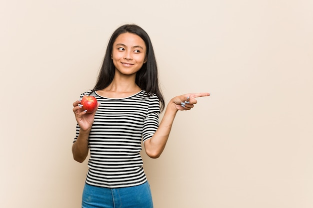 Mulher asiática nova que prende uma maçã que sorri e que aponta de lado, mostrando algo no espaço em branco.