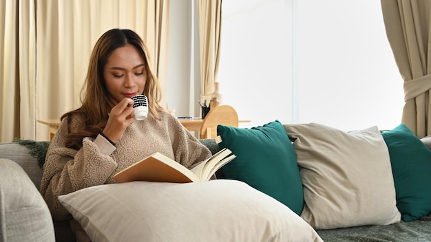 Mulher asiática pacífica lendo livro e tomando café no sofá