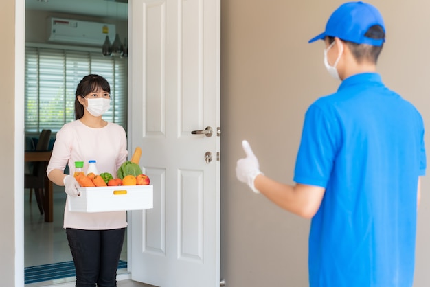 Mulher asiática pegar caixa de compras de alimentos, frutas, vegetais e bebidas e polegar para cima sem contato ou contato livre de entregador na frente da casa para distanciamento social para o risco de infecção.