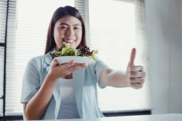 Mulher asiática polegares para cima e segurando o prato vegetal para uma dieta saudável com sorriso. Bela mulher sorridente comendo salada vegetariana orgânica fresca. Mulher jovem comendo salada saudável no conceito de dieta.