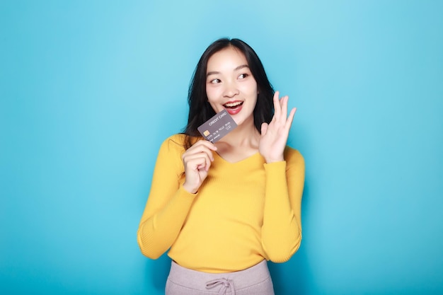 Mulher asiática posando com um cartão de crédito Retrato de uma bela jovem em um fundo azul claro feliz e sorridente posando em posição de pé