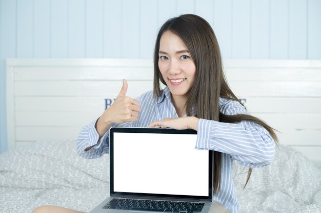 Mulher asiática que sorri e que mostra a tela de laptop vazia em seu quarto.