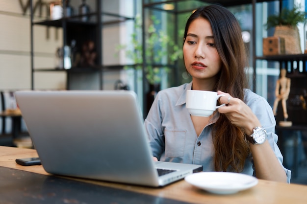 Mulher asiática que trabalha e bebe café no café com sorriso de computador portátil e trabalho feliz