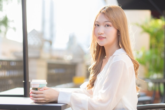 Mulher asiática que usa terno com cabelo cor de bronze se senta na cadeira de um café enquanto segura uma xícara de café