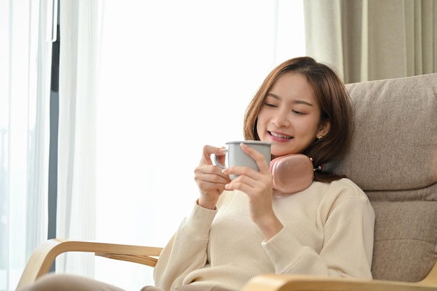 Mulher asiática relaxada e pacífica relaxando em sua poltrona confortável e tomando café na sala de estar