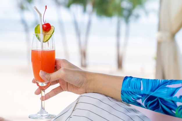 Mulher asiática relaxada na praia com seu coquetel de cereja e limão vermelho na mão na abertura do dia de verão