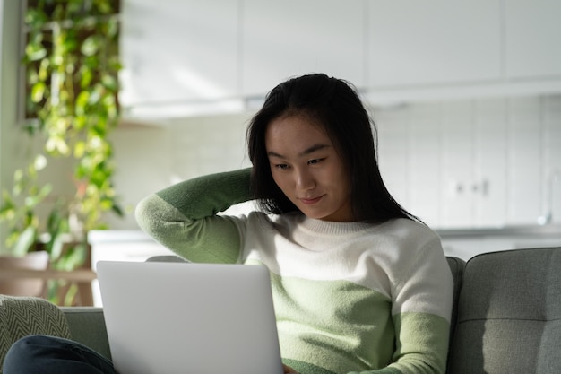 Mulher asiática relaxada positiva senta-se no sofá com o laptop de joelhos pensando em fazer compras na loja online