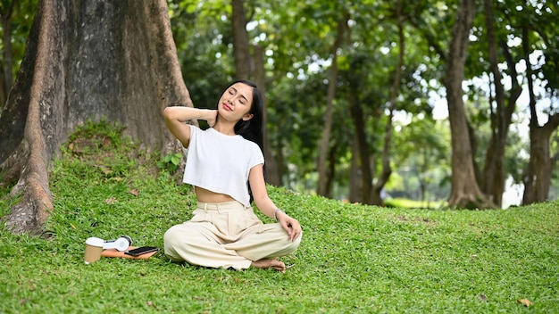 Mulher asiática relaxada sentada debaixo da árvore esticando e massageando o pescoço
