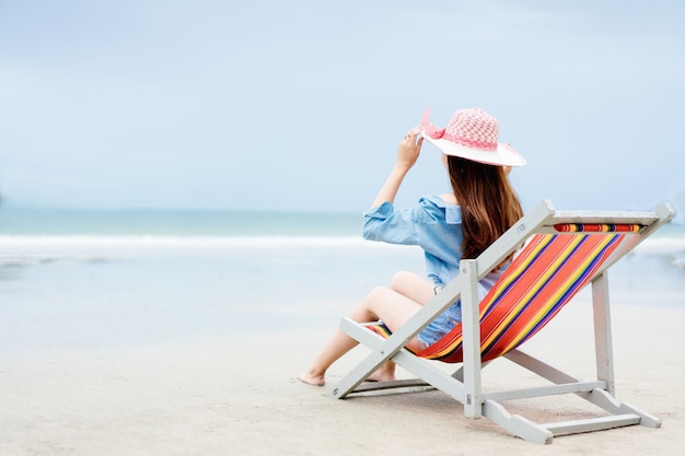 Mulher asiática relaxando em uma cadeira de praia e levantando os braços para pegar um chapéu