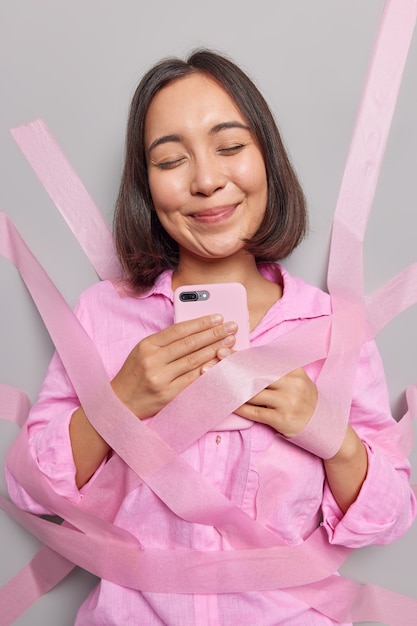 Mulher asiática satisfeita segura celular usa aplicativo moderno satisfeita em receber mensagem de bate-papos de namorado nas redes sociais fecha os olhos de prazer embrulhado em fitas veste camisa rosa