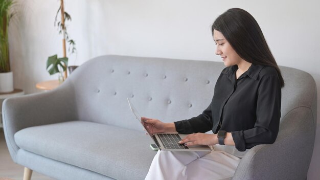 Mulher asiática satisfeita sentada no sofá e usando laptop para navegar na internet