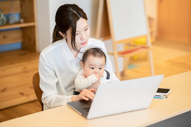 Mulher asiática segurando um bebê e operando um laptop dentro de casa