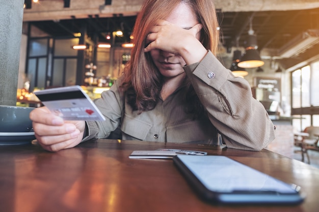 Foto mulher asiática segurando um cartão de crédito, sentindo-se estressada e sem dinheiro, celular na mesa