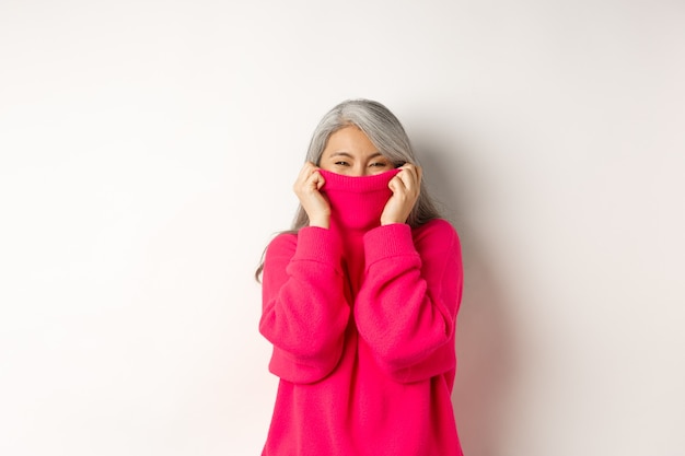 Mulher asiática sênior alegre rindo, vestindo um suéter quente, escondendo o rosto dentro da gola, feliz em pé sobre um fundo branco.