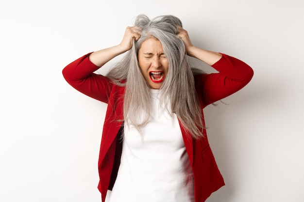 Mulher asiática sênior angustiada e irritada puxando o cabelo e gritando, em pé chateada com um fundo branco