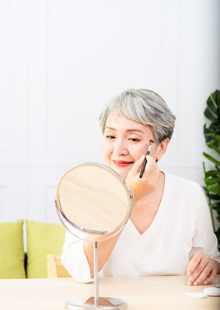 Mulher asiática sênior aplicando base na bochecha com um pincel de maquiagem enquanto está sentada sozinha na frente de um espelho.
