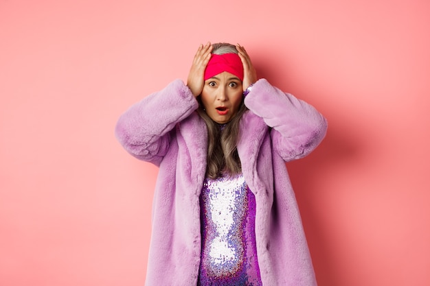 Mulher asiática sênior chocada e assustada, vestindo um casaco de pele artificial roxo, segurando as mãos na cabeça e em pânico, em pé angustiada contra um fundo rosa