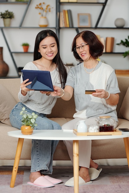 Mulher asiática sênior feliz e sua filha adulta aproveitando o tempo juntas, elas estão bebendo chá e fazendo compras online