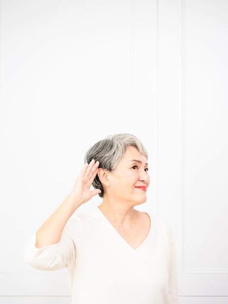 Mulher asiática sênior sorrindo com a mão na orelha, ouvindo uma audiência de boatos ou fofocas.